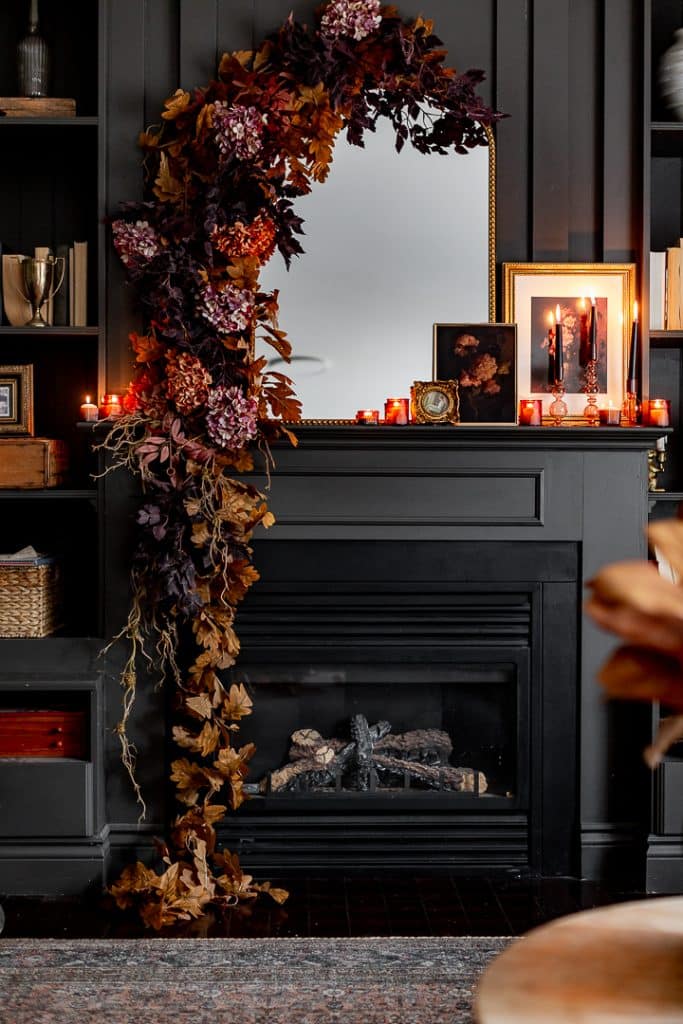 A dark, elegant fireplace is decorated with cascading autumn leaves and flowers around a mirror. A Dough Bowl Candle and framed pictures sit on the mantel, creating a warm, cozy atmosphere against dark paneled walls.