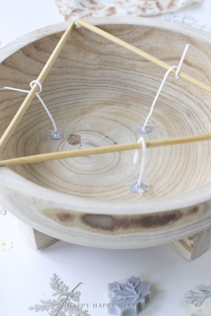 A wooden dough bowl candle DIY features three wooden sticks arranged in a triangle across the top, secured by white cords and glue at the ends inside the bowl.