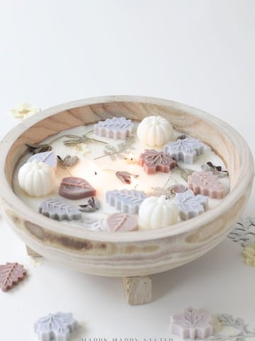 A wooden dough bowl candle diy adorned with small, colorful wax pumpkins and leaf shapes, surrounded by dried flowers, creates a cozy autumn-inspired centerpiece.