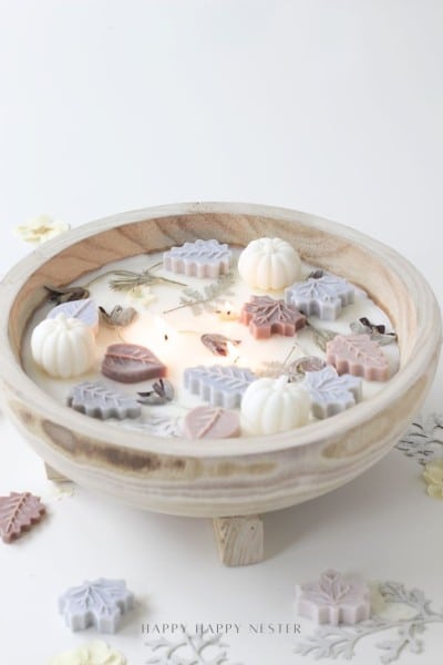 A wooden dough bowl candle diy adorned with small, colorful wax pumpkins and leaf shapes, surrounded by dried flowers, creates a cozy autumn-inspired centerpiece.