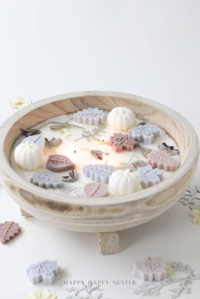 A wooden bowl candle inspired by all things pumpkin, decorated with wax shapes of leaves and pumpkins in pastel colors. Dried flowers are scattered on creamy white wax, illuminated by a soft flame in the center.