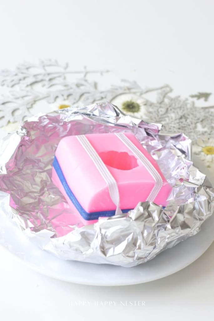 A pink rectangular ice cream sandwich mold with a heart-shaped cutout sits on a plate, partially wrapped in crinkled aluminum foil—an inspiring scene for any dough bowl candle DIY enthusiast. White flowers are visible in the background.