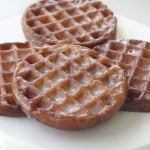 Three round glazed donuts and two rectangular glazed donuts with a waffle-like pattern are arranged on a white surface.