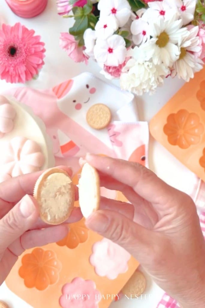 A person opens a cream-filled cookie in a bright, cheerful setting with pink and white flowers, floral molds, and cookies arranged on a table.