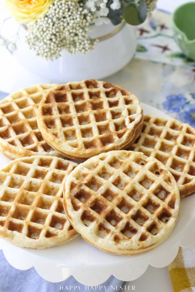 Five golden-brown waffles are stacked on a white cake stand, with a floral arrangement and a green cup in the background. The scene looks bright and inviting, evoking a cheerful breakfast setting.