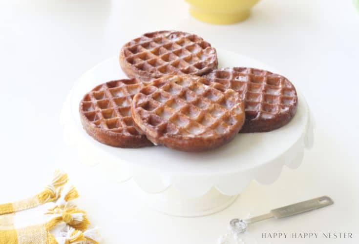 Four glazed waffle donuts are arranged on a white scalloped cake stand. A yellow and white cloth and a measuring spoon sit nearby on a bright, white surface.