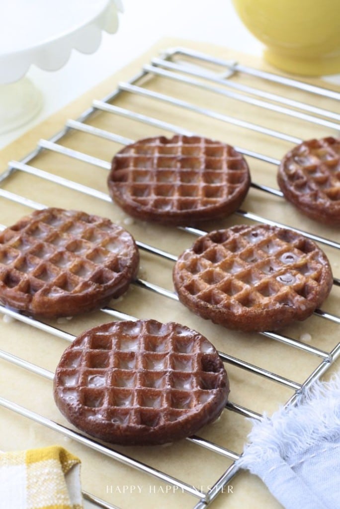 Five round, golden-brown waffles with a shiny glaze are cooling on a metal rack. The background includes a yellow cup, a white plate, and a blue cloth.