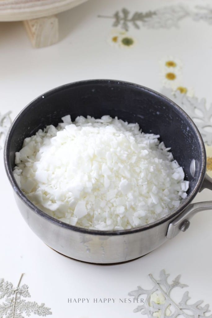 A black saucepan filled with white wax flakes sits on a white surface decorated with pressed flowers and leaves, ready for your next dough bowl candle DIY project.