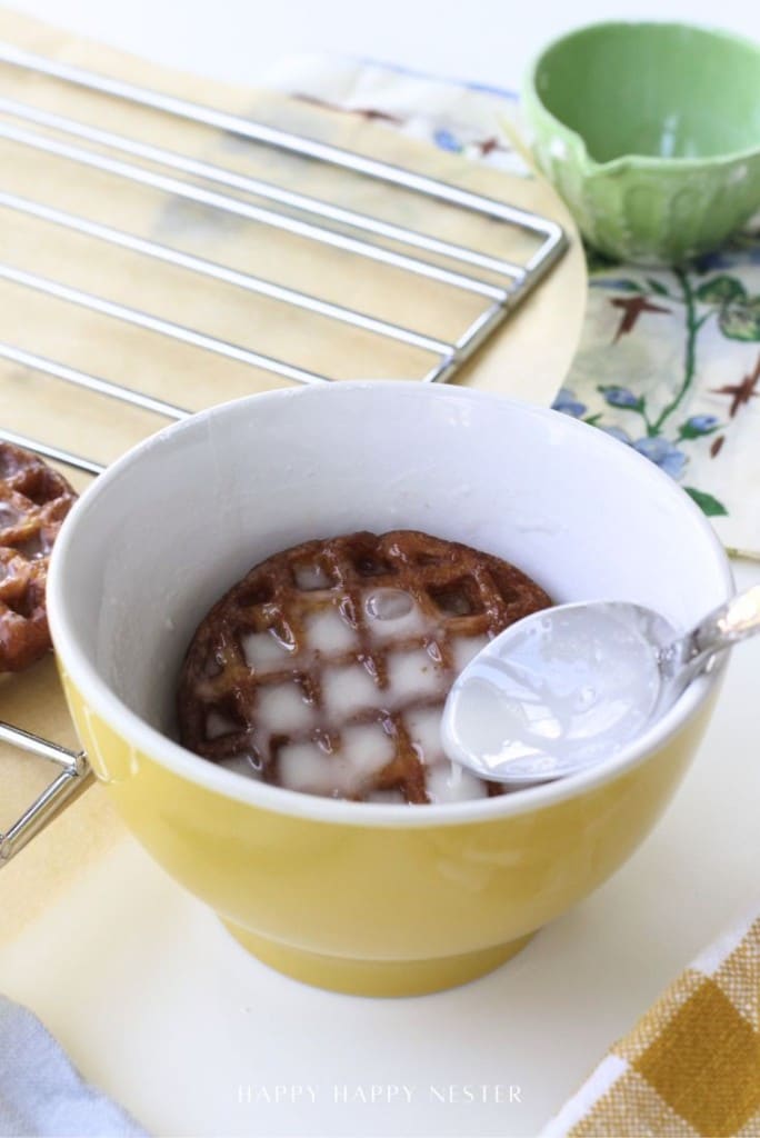 A waffle cookie, inspired by our mini waffle donuts recipe, sits in a yellow bowl as white icing is drizzled on top from a spoon. A cooling rack and a green bowl rest in the background on a patterned cloth.