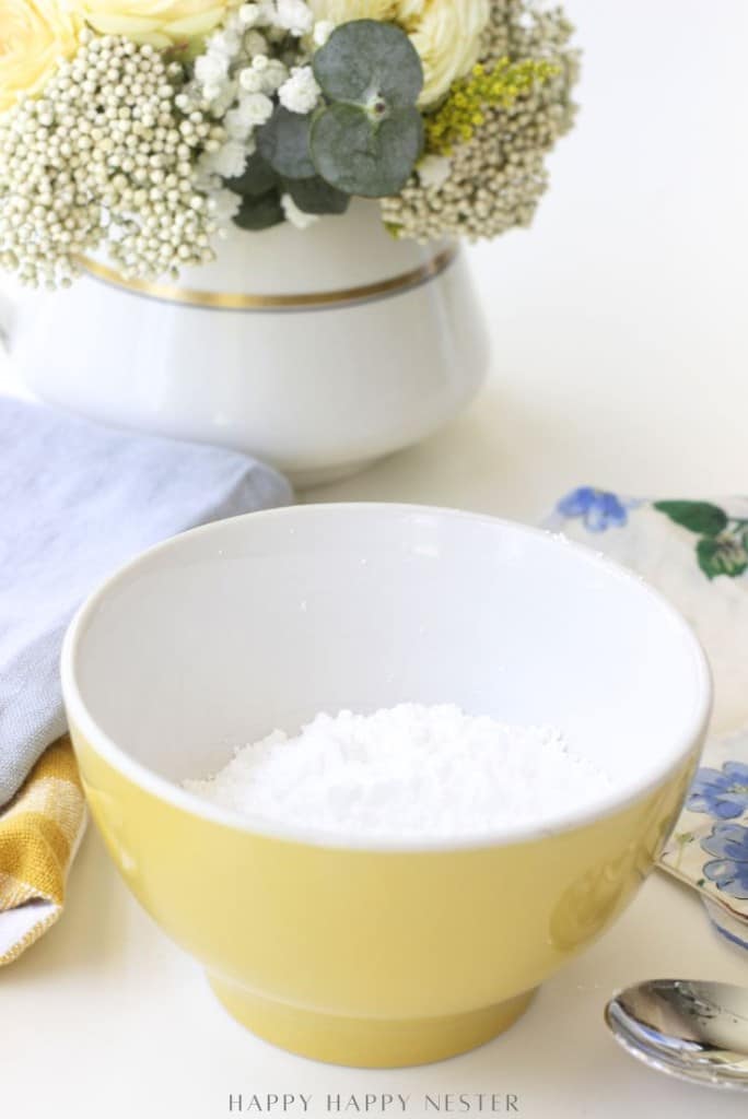 A yellow bowl filled with powdered sugar sits on a table beside a blue napkin, a spoon, and a floral arrangement in a white vase—perfect for dusting your favorite mini waffle donuts recipe.