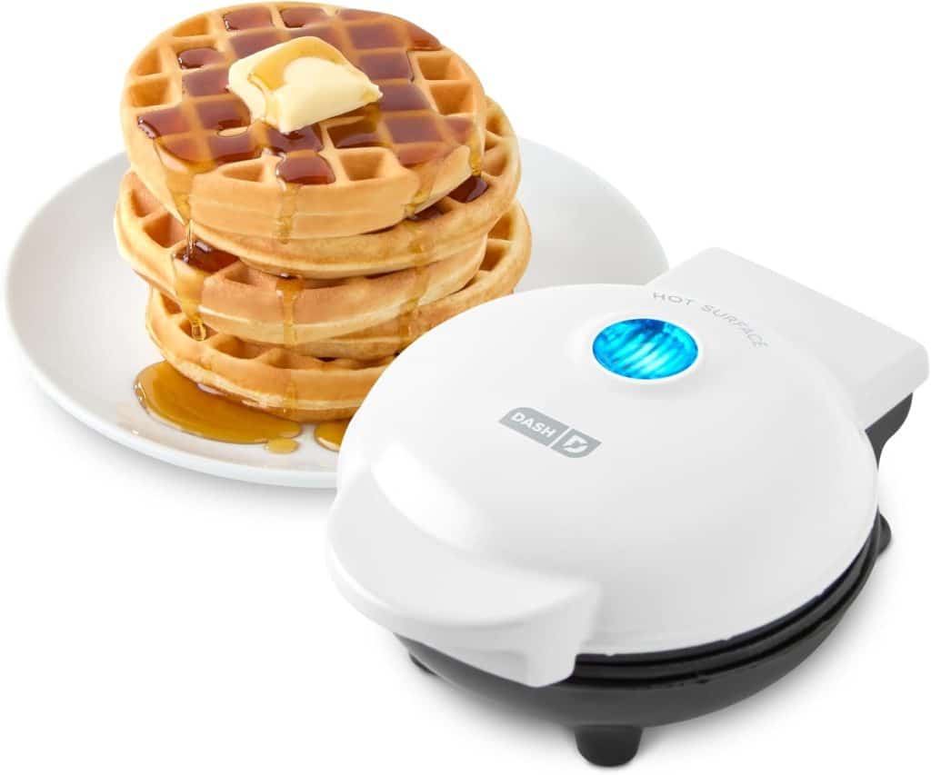 A small white waffle maker sits next to a plate stacked with three waffles topped with butter and syrup, perfect for enjoying alongside your favorite Mini Waffle Donuts Recipe, with syrup drizzling down the sides.