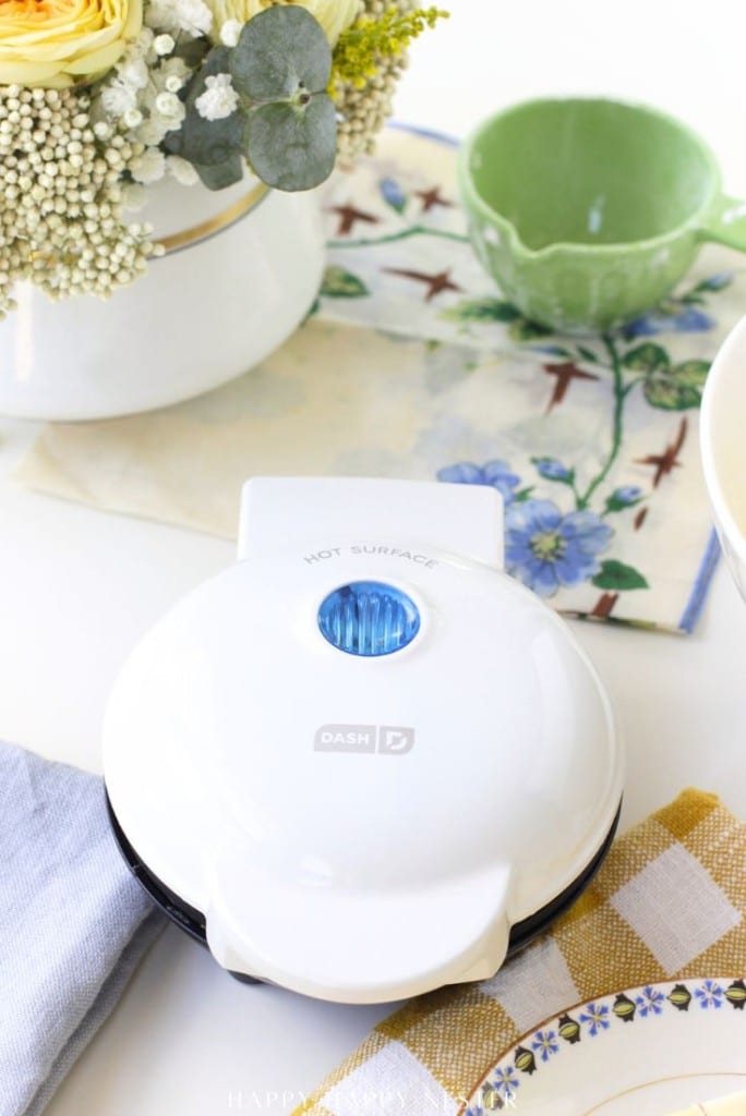 A white Dash mini waffle maker with a blue indicator light sits on a table next to a floral arrangement, a green measuring cup, and a blue napkin—perfect for trying out your favorite mini waffle donuts recipe.