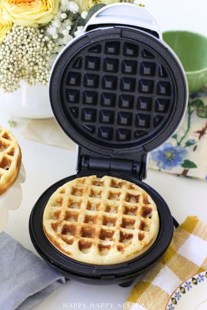 A golden waffle, inspired by a mini waffle donuts recipe, sits inside an open waffle maker on a kitchen counter, with a floral arrangement, green cup, and patterned napkin in the background.