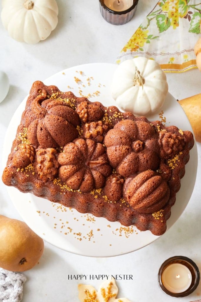 A rectangular pumpkin spice loaf cake, perfect for fans of all things pumpkin, is decorated with raised pumpkin and leaf shapes and gold sprinkles. It sits on a white cake stand surrounded by candles, white mini pumpkins, and pears.