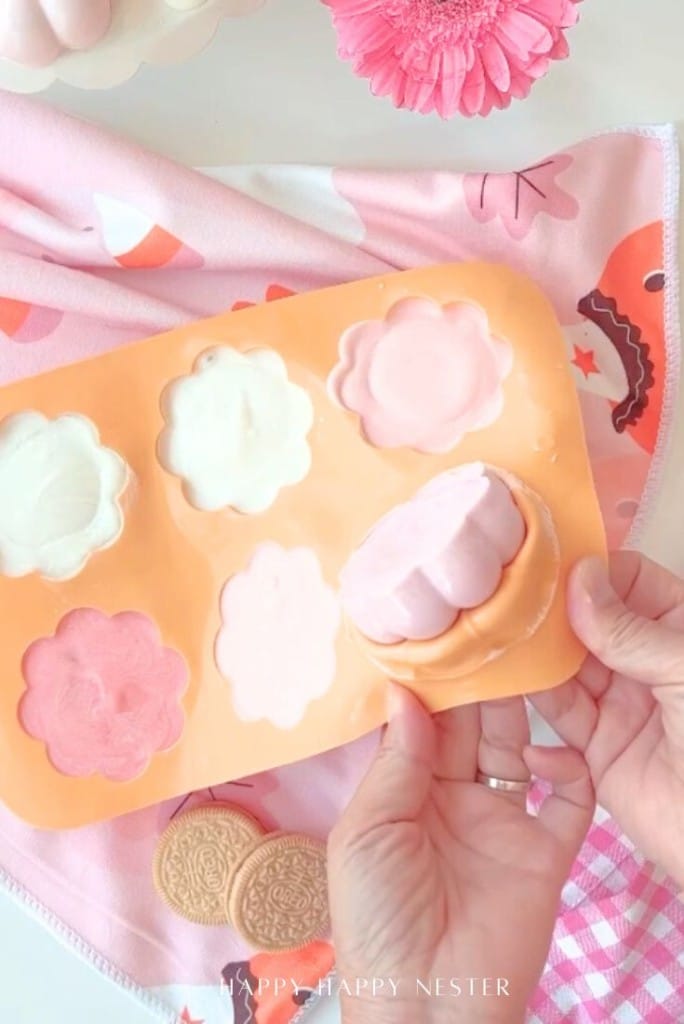 A hand presses a pink, flower-shaped treat from a silicone mold with other pastel-colored flower treats nearby, on a pink napkin with a fox print and some vanilla sandwich cookies—perfect for an Oreo Halloween treats recipe.