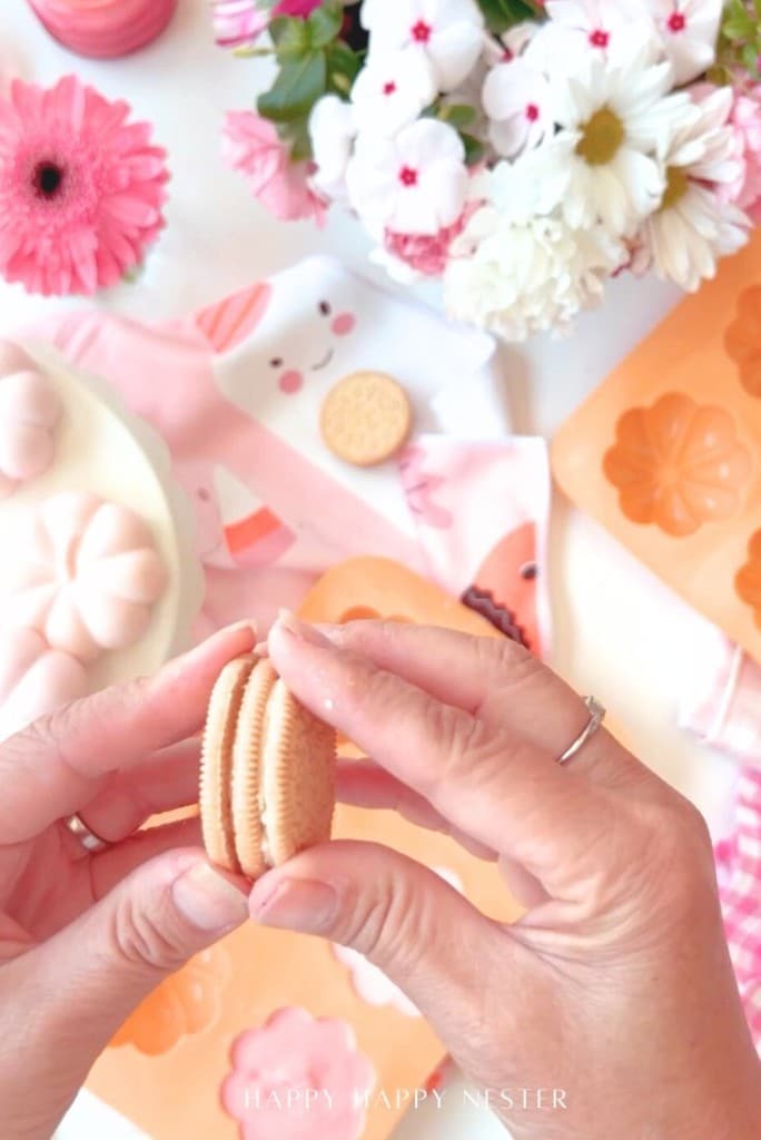 Hands holding a sandwich cookie above a light pink surface with floral napkins, flowers, and cookie molds. Other cookies, a pink flower, and baking molds are visible in the background.