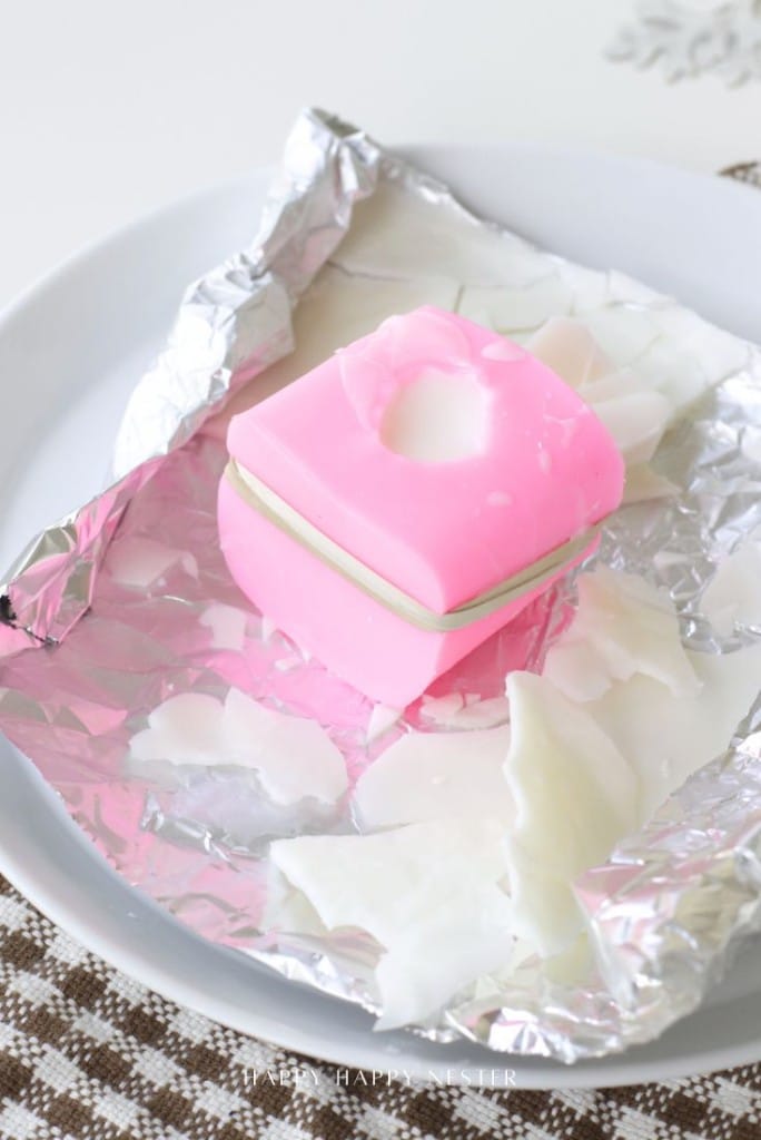 A pink square soap with a circular indentation sits on aluminum foil, surrounded by white soap shavings—perfect inspiration for a dough bowl candle DIY—on a white plate atop a brown checkered cloth.