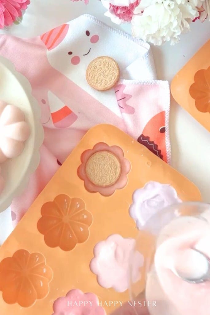 Pink and white flower-shaped treats in a silicone mold, a vanilla sandwich cookie, a cute napkin with a smiling design, and pink flowers create a cheerful, pastel scene.