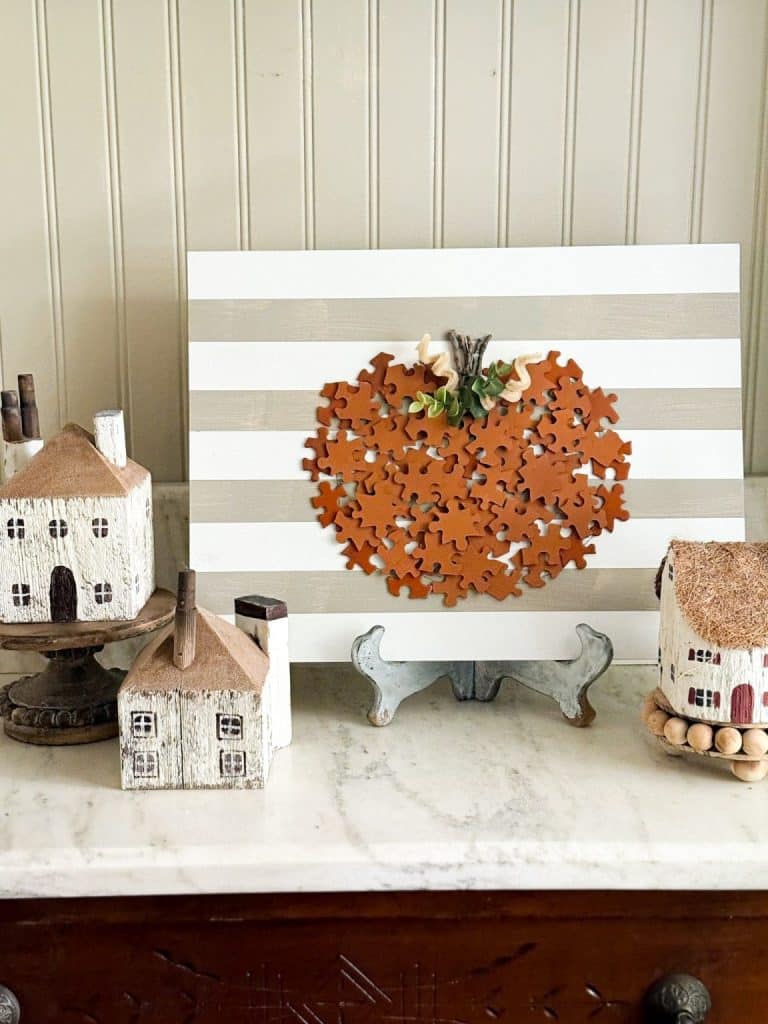 A striped white and beige sign with an orange pumpkin made from puzzle pieces celebrates all things pumpkin, displayed on a stand and surrounded by small decorative house figurines on a marble surface.