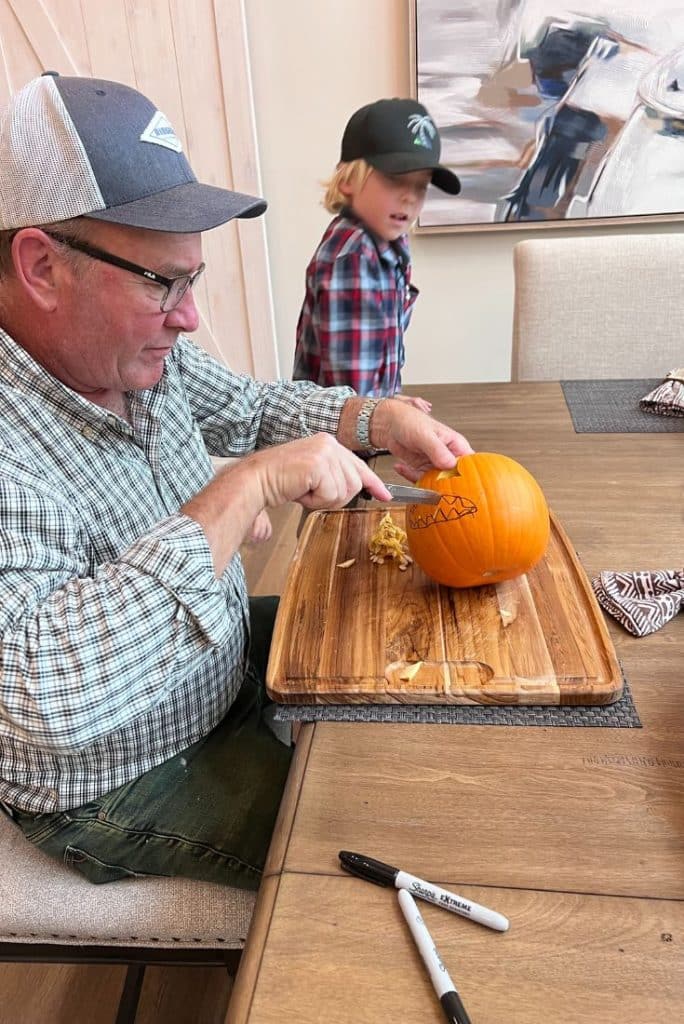A man wearing glasses and a cap carves a pumpkin on a wooden cutting board at a table, celebrating all things pumpkin, while a young blonde child in a plaid shirt and cap stands in the background. Two markers and napkins are on the table.