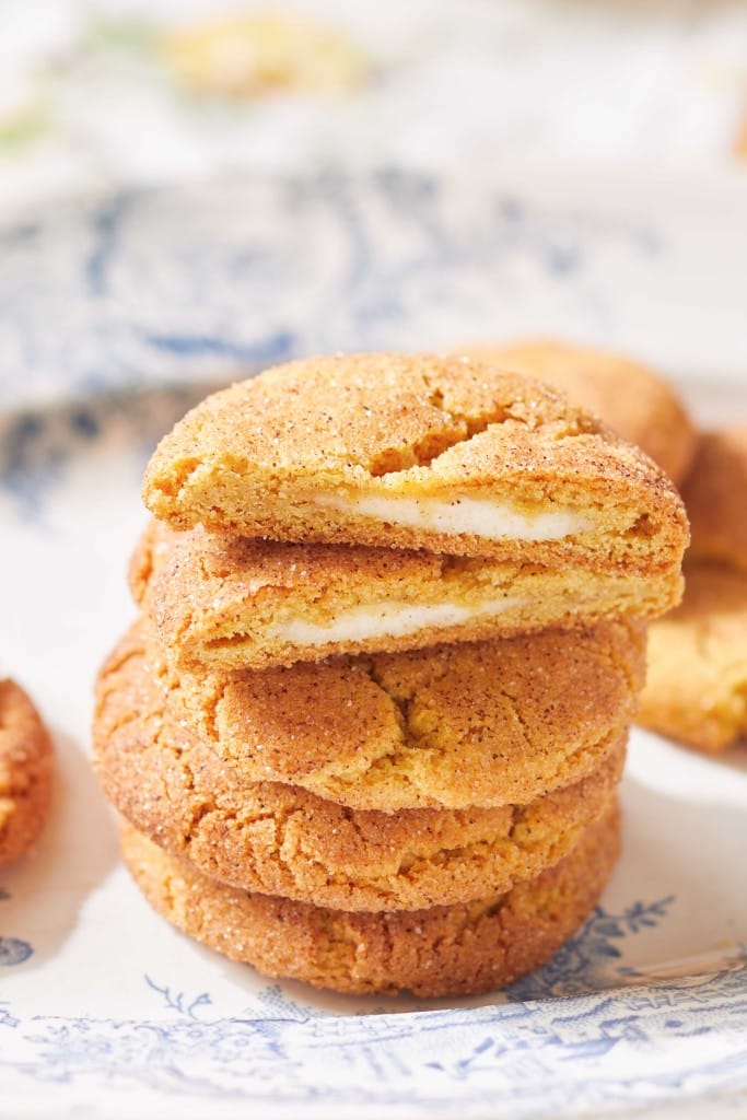 A stack of golden brown cookies with cinnamon sugar coating, one broken in half to reveal a creamy white filling, hints at all things pumpkin, displayed on a decorative white and blue plate.