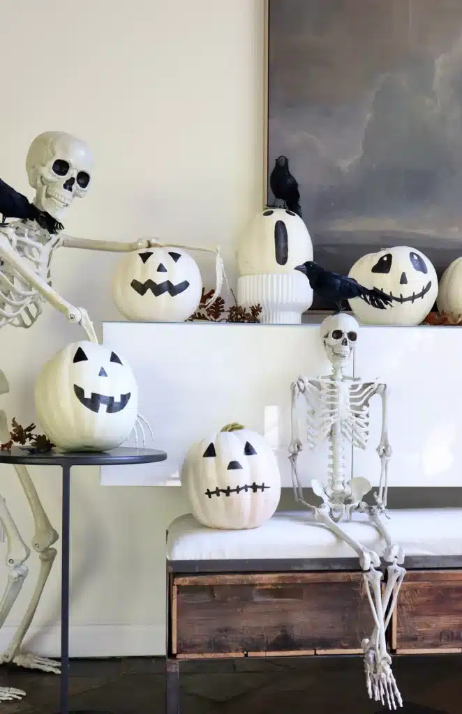 Two skeleton figures pose with white pumpkin crafts painted as jack-o’-lanterns, while black faux crows perch nearby, creating a playful Halloween display indoors.