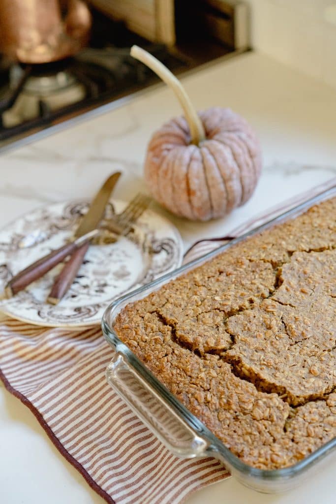 A glass baking dish filled with pumpkin bread pudding sits on a striped cloth next to a vintage plate with utensils and a decorative fabric pumpkin on a marble kitchen counter, perfect for trying a cozy pumpkin bread pudding recipe.