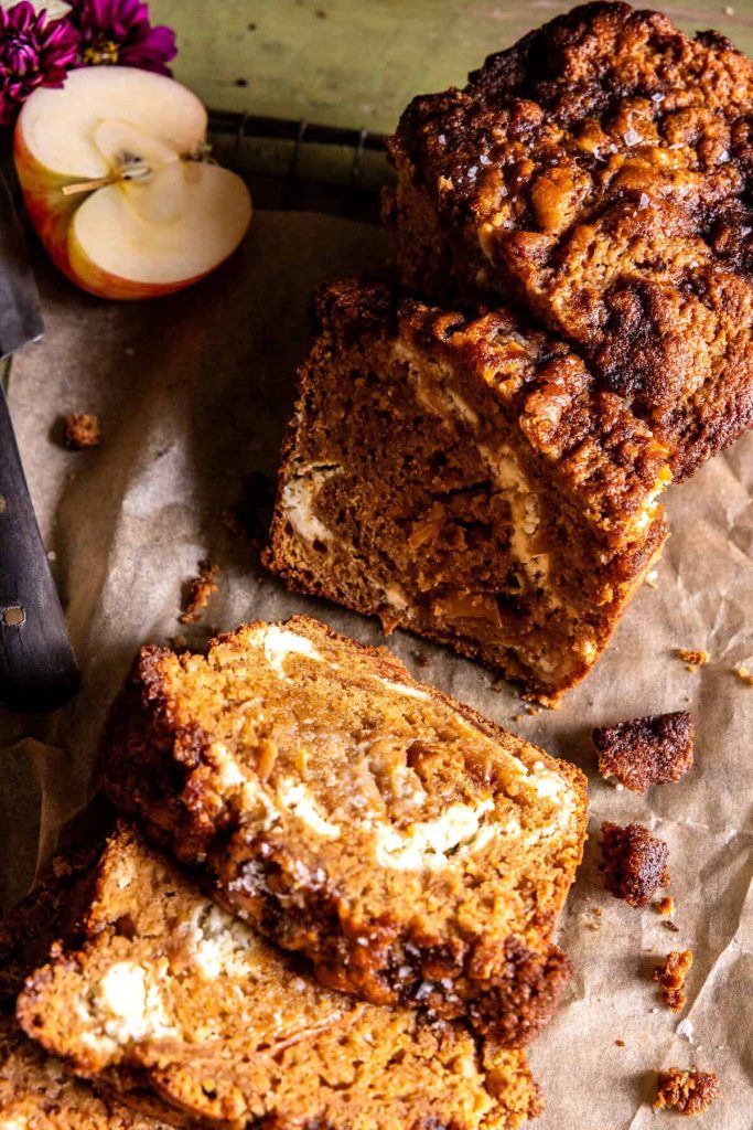 A sliced loaf of apple bread with visible chunks of apple and swirls of cream cheese on parchment paper, next to a knife and a halved apple—perfect for homemade Thanksgiving treats.