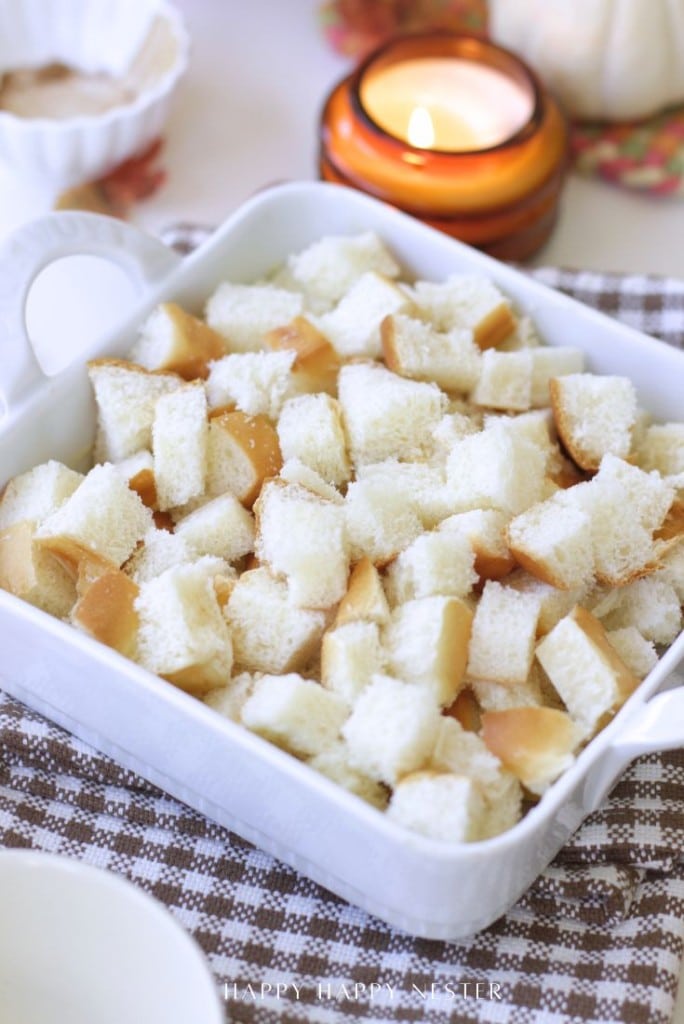 A white ceramic baking dish filled with cubes of white bread, some with crusts, sits on a checkered cloth—perfect for trying a pumpkin bread pudding recipe. A lit candle and white bowl are in the blurred background.
