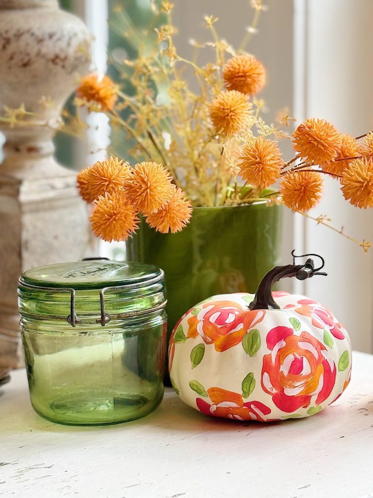 A painted pumpkin with colorful floral designs sits on a white surface beside a green glass jar and a green vase filled with dried orange flowers—my favorite Thanksgiving decor, glowing as sunlight filters in from the background.