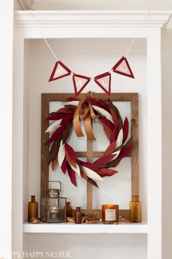 A fall-themed shelf display features a red, brown, and white leaf wreath with a brown ribbon on a window frame, small vintage bottles, candles, leaves, a lantern, and wall crafts for fall like a red triangle pennant garland hanging above.