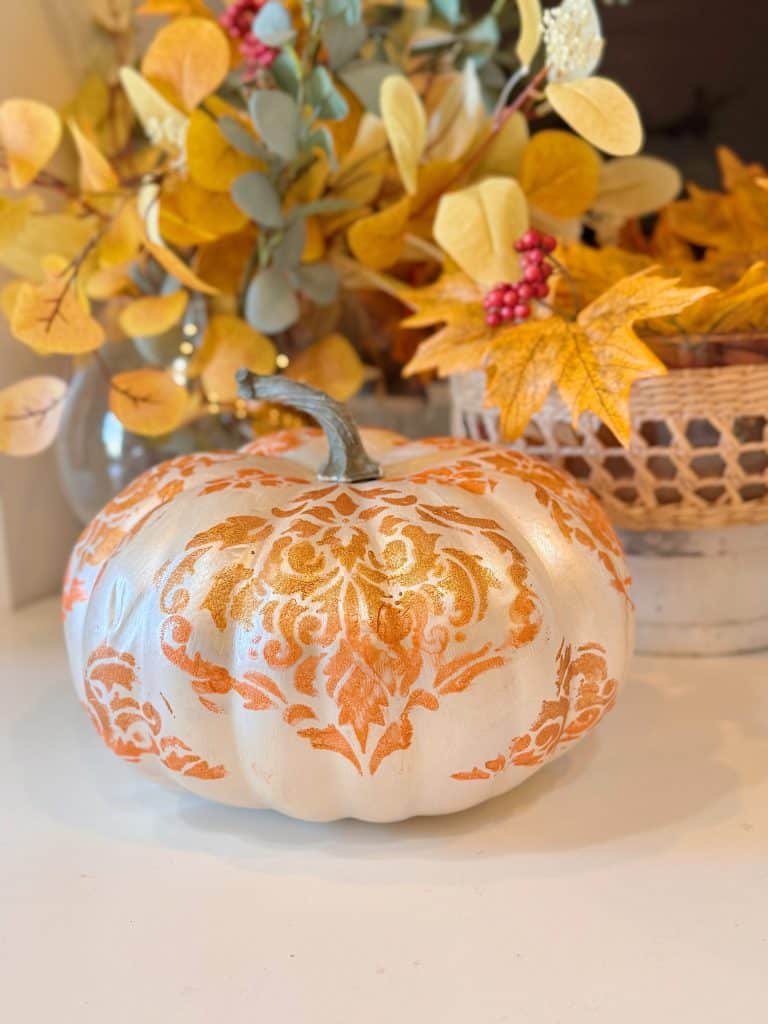 A white pumpkin with intricate orange patterns, created using temporary tattoos, sits among autumn leaves, berries, and a wicker basket—one of the easiest no carve pumpkins and stylish pumpkin decorating ideas for fall.