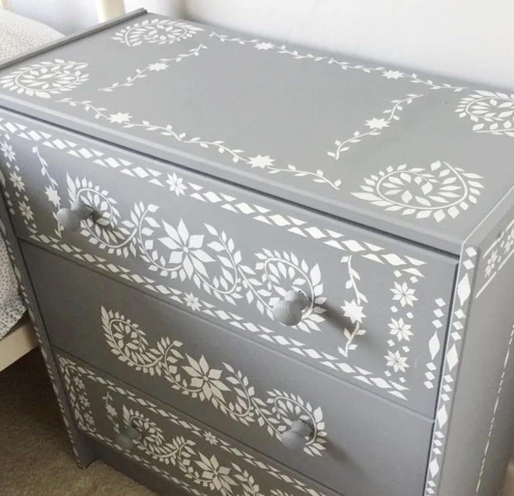 A gray dresser with three drawers, accented by intricate white floral and geometric patterns reminiscent of white pumpkin crafts. Each drawer features two round knobs, adding a charming, handcrafted touch.