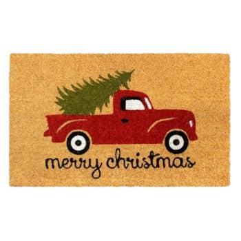 A doormat with a red vintage truck carrying a Christmas tree and the words "merry christmas" written below on a tan background.