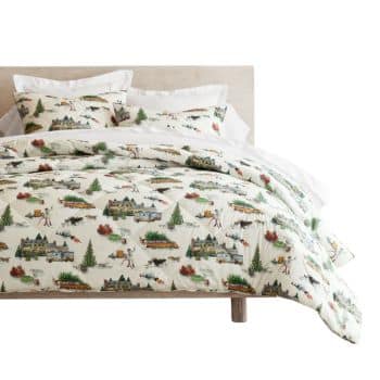 A neatly made bed with a light wood headboard features bedding decorated with colorful illustrations of houses, trees, people, and animals on a white background. Matching pillows and comforter complete the look.