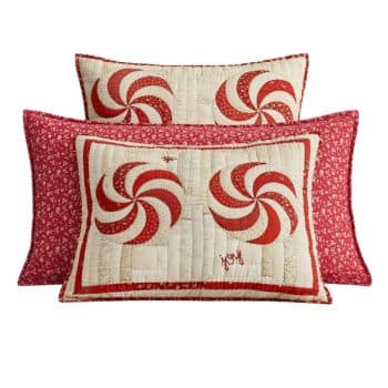 Three decorative pillows with red and white peppermint swirl patterns and red floral borders. One pillow has the word "joy" stitched in red in the lower right corner.