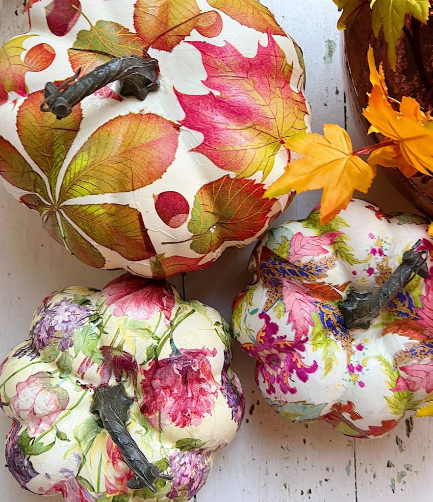 Three decorative pumpkins, including white pumpkin crafts covered in colorful, floral and leaf-patterned designs, are arranged on a white surface, with a few artificial autumn leaves nearby.