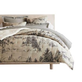 A neatly made bed with layered pillows and a comforter featuring a forest and mountain print in neutral colors, set against a light wooden headboard.