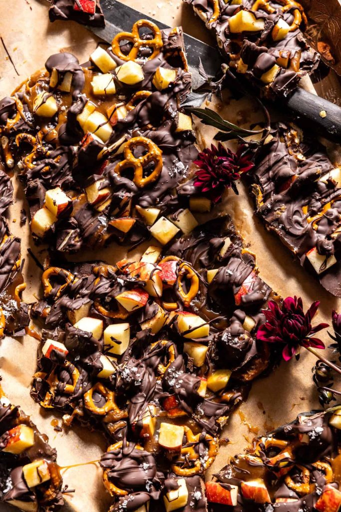 Chocolate bark topped with pretzels, diced apples, and a drizzle of dark chocolate is scattered on parchment paper, accented with red flowers—perfect for festive Halloween treats.