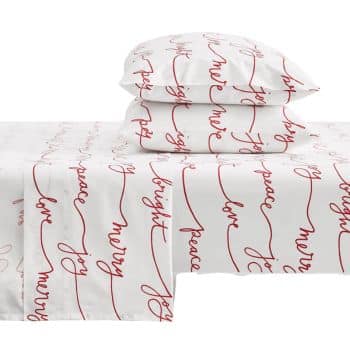 White bed sheets and pillowcases with red cursive words like "merry," "joy," "peace," "bright," and "love" repeating in horizontal lines as a festive holiday-themed design.