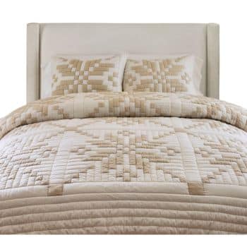 A neatly made bed with a white upholstered headboard, featuring a quilt and matching pillows in beige and cream tones with a geometric, patchwork star pattern.