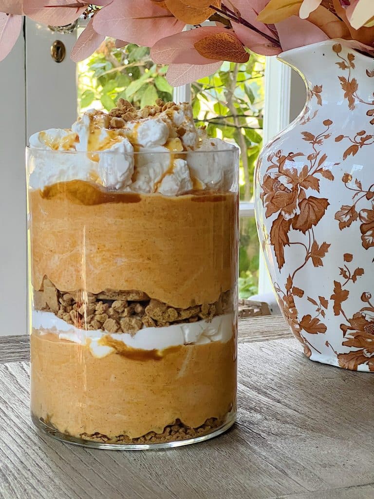A glass filled with layers of creamy pumpkin mousse, crumbled graham crackers, whipped cream, and caramel sauce sits on a wooden table beside a decorative white vase—perfect for festive Thanksgiving treats.