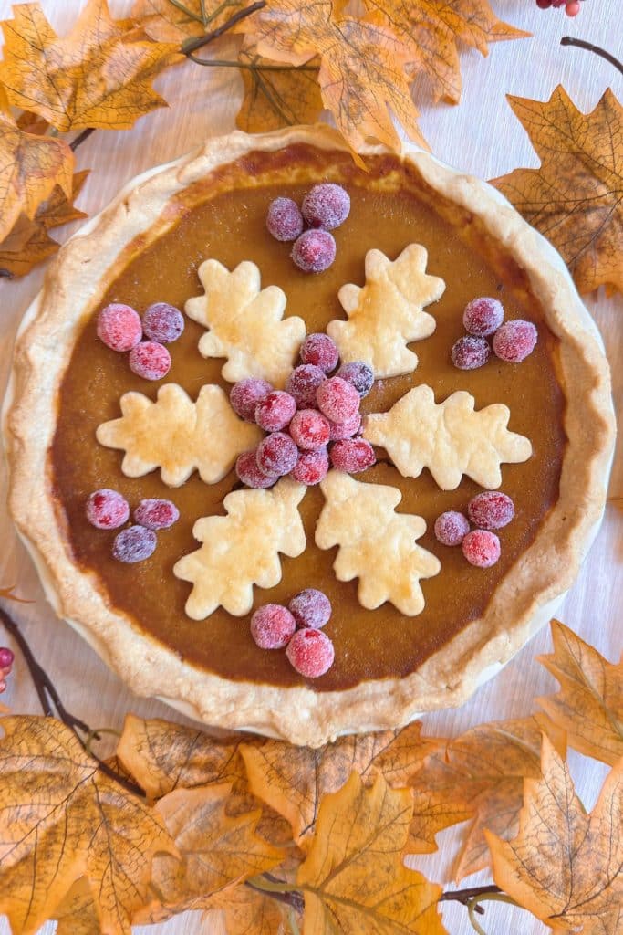 Pumpkin pie decorated with leaf-shaped pastry cutouts and sugared cranberries, surrounded by artificial autumn leaves on a light surface—a cozy scene perfect for pairing with your favorite pumpkin bread pudding recipe.