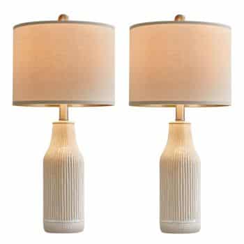 A pair of matching table lamps with ribbed, beige ceramic bases and large, cylindrical beige fabric lampshades, both turned on and emitting a soft, warm light.