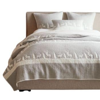A neatly made bed with a beige headboard, featuring a light beige and white quilt and matching pillows decorated with silhouettes of reindeer and trees in a winter forest design.