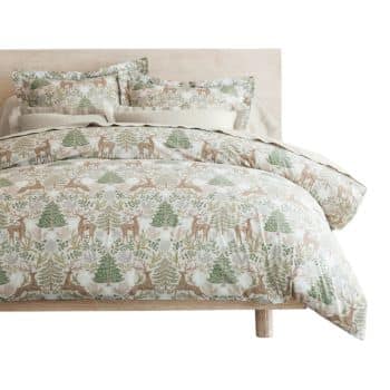 A neatly made bed with a light wood frame, featuring a comforter and pillows decorated with a woodland pattern of green trees and brown deer on a cream background.
