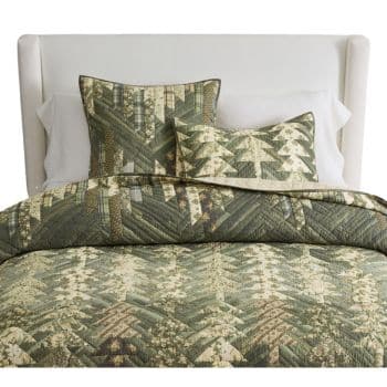 A bed with a white upholstered headboard, covered by a green and beige patchwork quilt with a tree pattern, and matching pillow shams.