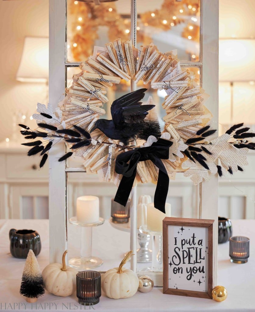 A Halloween-themed table display with a wreath made of book pages, perfect for wall crafts for fall, features a black crow, candles, small pumpkins, and a sign that reads "I put a spell on you.