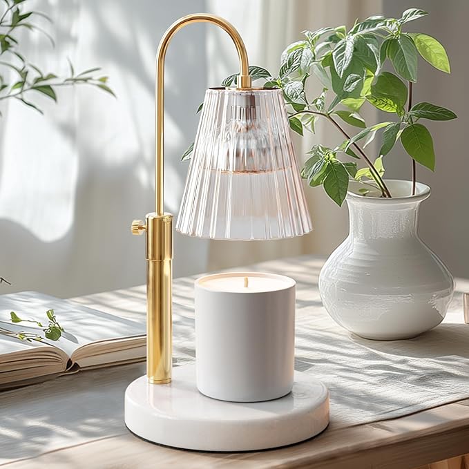 A gold and glass candle warmer lamp—my favorite Thanksgiving decor—stands on a marble base beside a white ceramic vase with green leaves, an open book, and sunlight streaming through the window in the background.