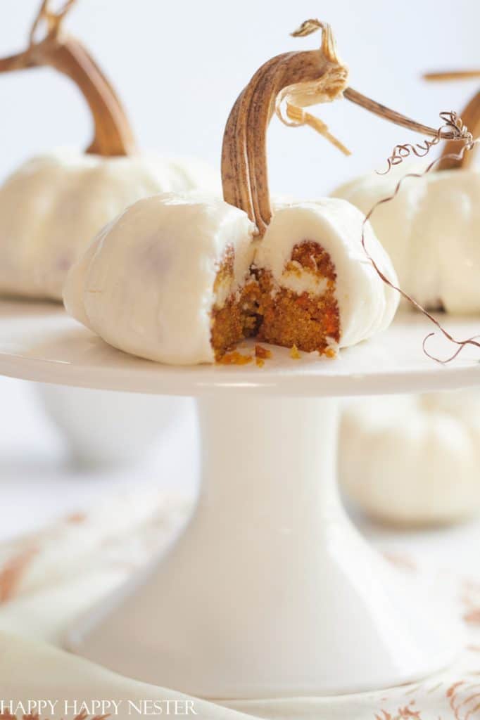 A pumpkin-shaped mini cake, perfect for halloween treats, is coated in white icing with a real pumpkin stem on top. The cake sits on a stand, cut open to reveal its moist orange interior and creamy filling.